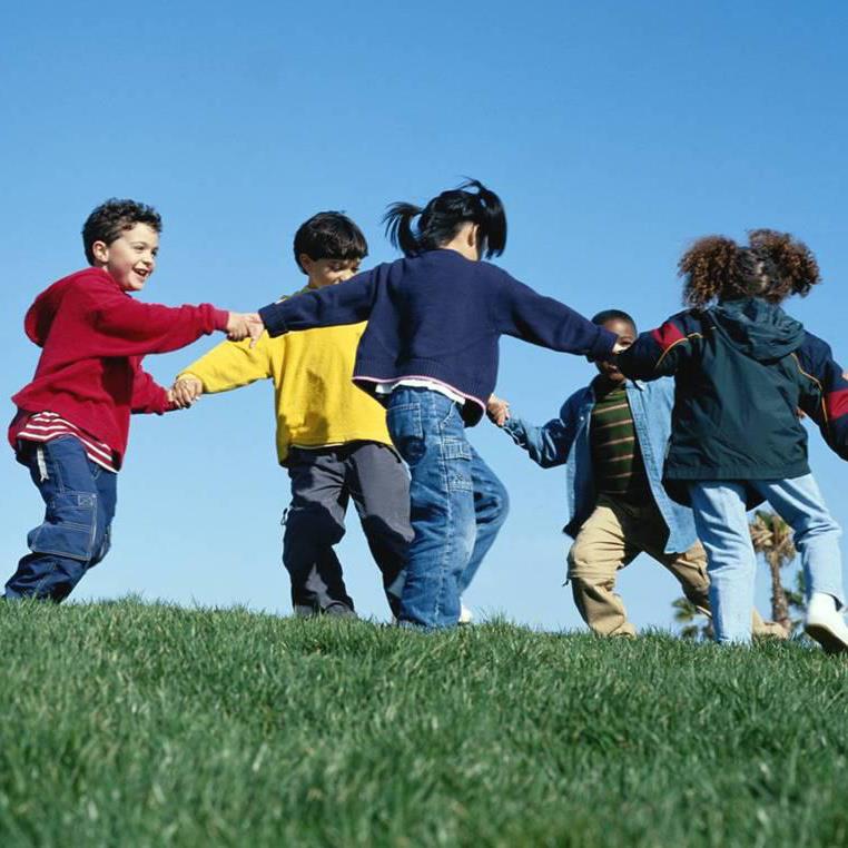 Multi-cultural children hold hands in a circle outside on green grass with a blue sky above them.