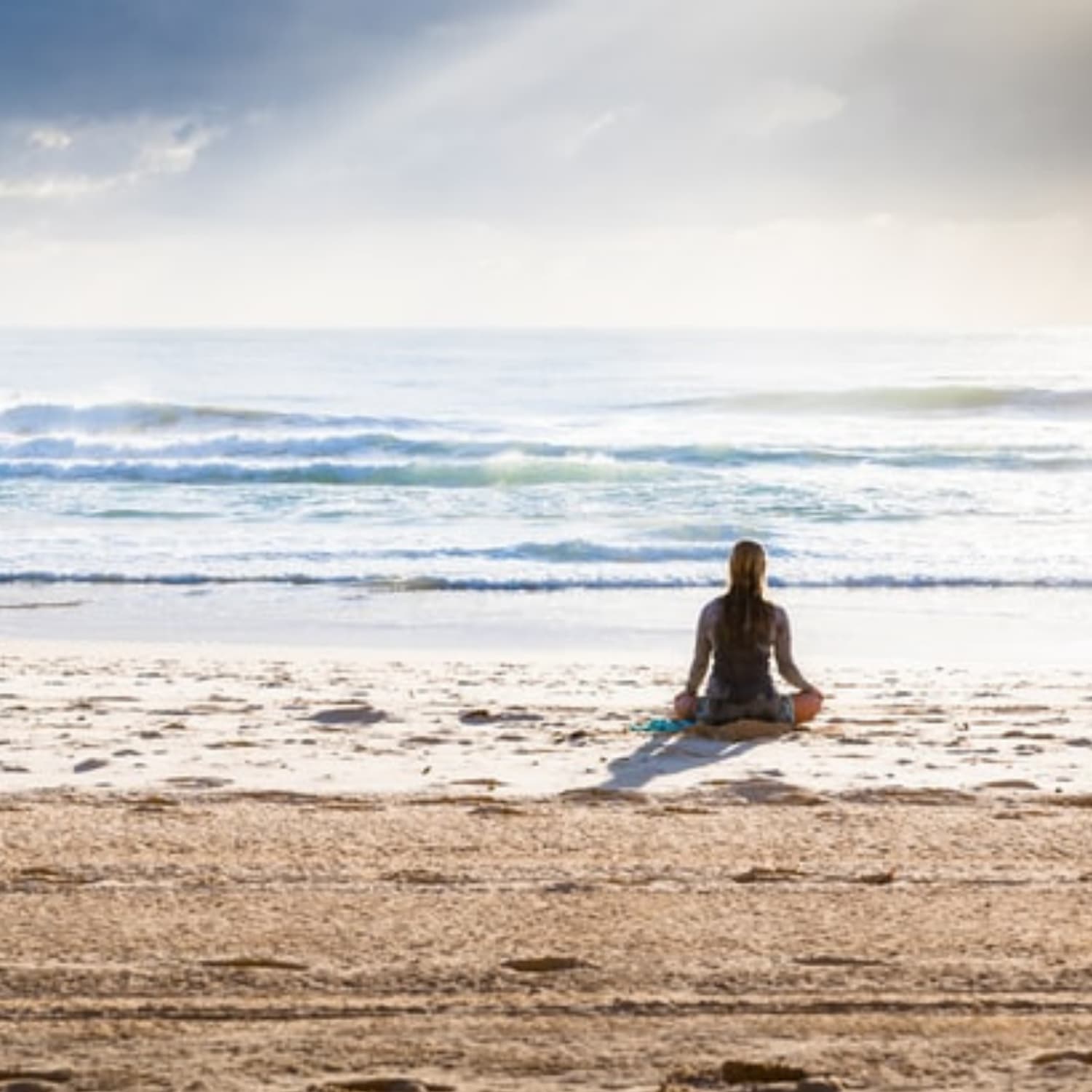 Woman seated cross-legged on a beach facing away from the camera.